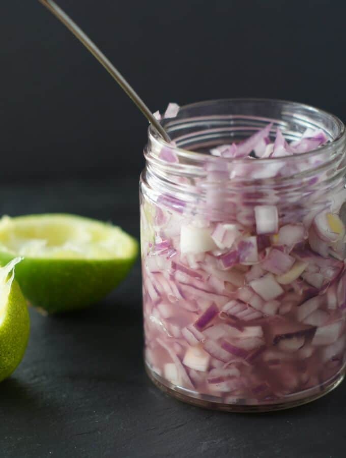 Lime marinated red onions in a glass jar with limes in the background on a black slate board