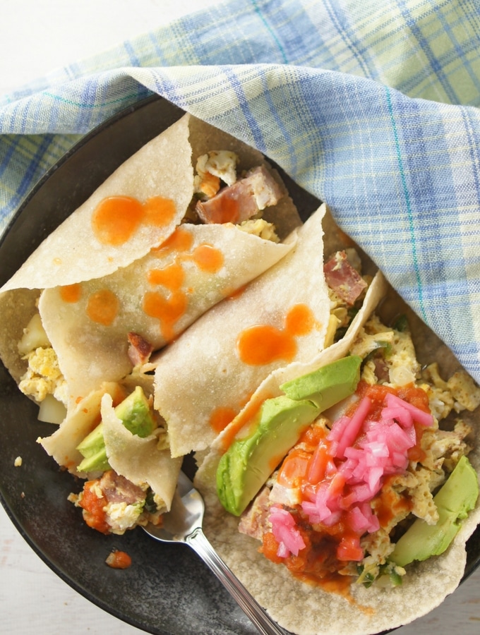 Three breakfast burritos with eggs, potatoes avocado and hot sauce over the top.